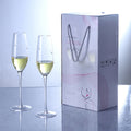 Two champagne flutes with white wine next to a branded gift box on a reflective surface.