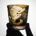 Decorative glass with gold animal engraving held by a gloved hand on a white background
