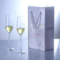 Two champagne flutes with white wine next to a branded gift box on a reflective surface.